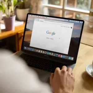 An adult using a laptop indoors, browsing Google at a wooden table with coffee.