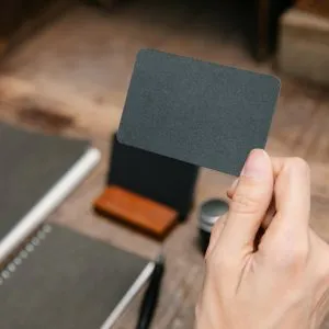 Close-up of a hand holding a black blank card with craft supplies in the background, offering ample copy space.